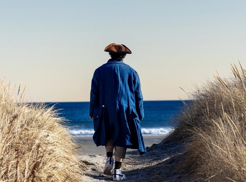 Person in colonial-style clothing walking toward the ocean between sand dunes.